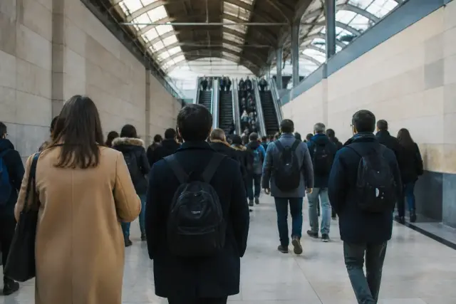 Passage couvert d’une grande gare, escaliers mécaniques et panneaux directionnels, voyageurs de dos en mouvement, éclairage de fin de journée