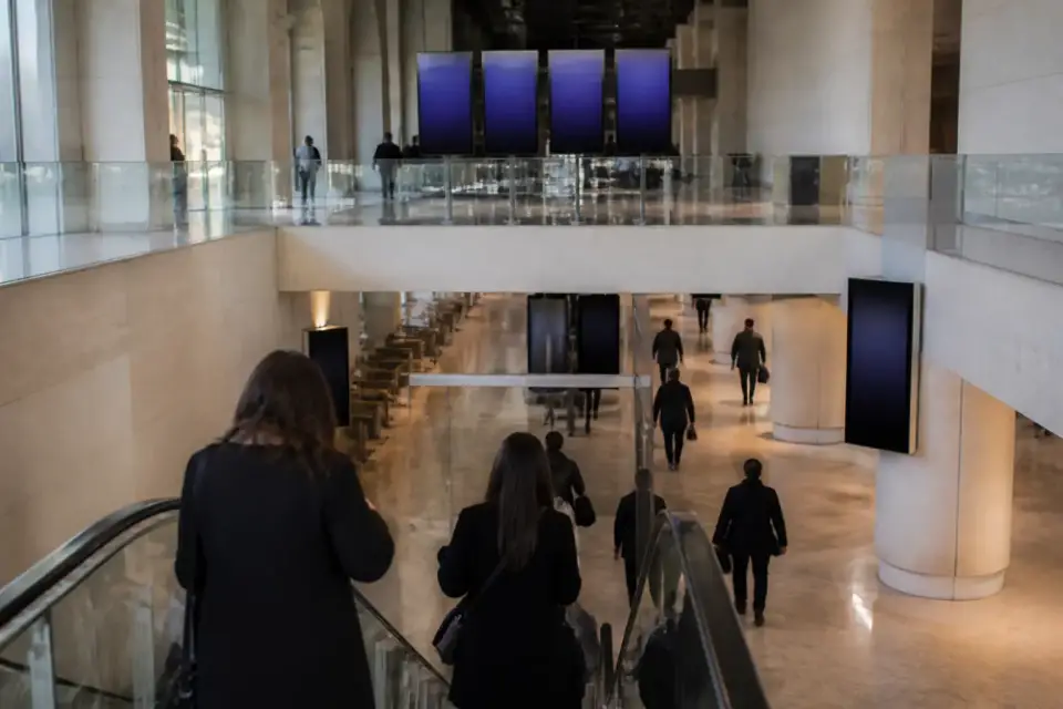 Hall d’aérogare sur deux niveaux, escalators et mezzanine, voyageurs de dos devant un tableau d’affichage vide, lumière vive