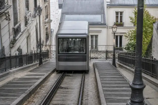 Une cabine de funiculaire vitrée sur une voie en pente entre deux escaliers de pierre, lumière du matin, sans personne ni inscription