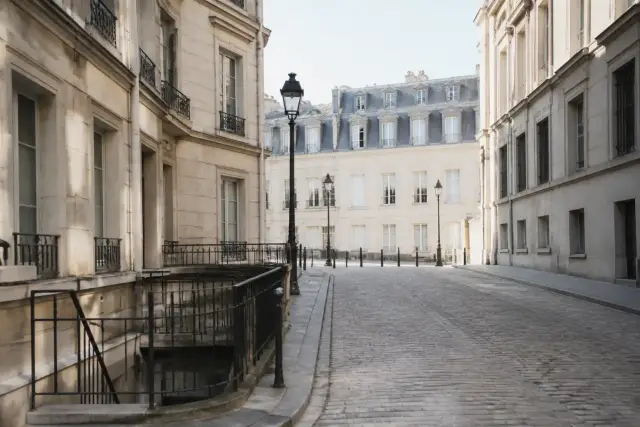 Une vaste cour dallée entourée de longs bâtiments de pierre claire sous un ciel dégagé, quelques silhouettes lointaines sans visage net