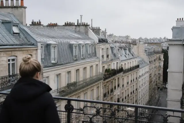 Pied d’une grande tour en fer à treillis vu d’en bas, jardins arborés au premier plan, ciel dégagé, visiteurs minuscules de dos