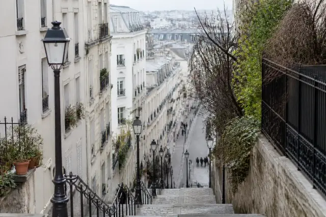 Une rue en pente bordée d’escaliers et de façades claires, lumière douce, quelques promeneurs lointains sans visage net